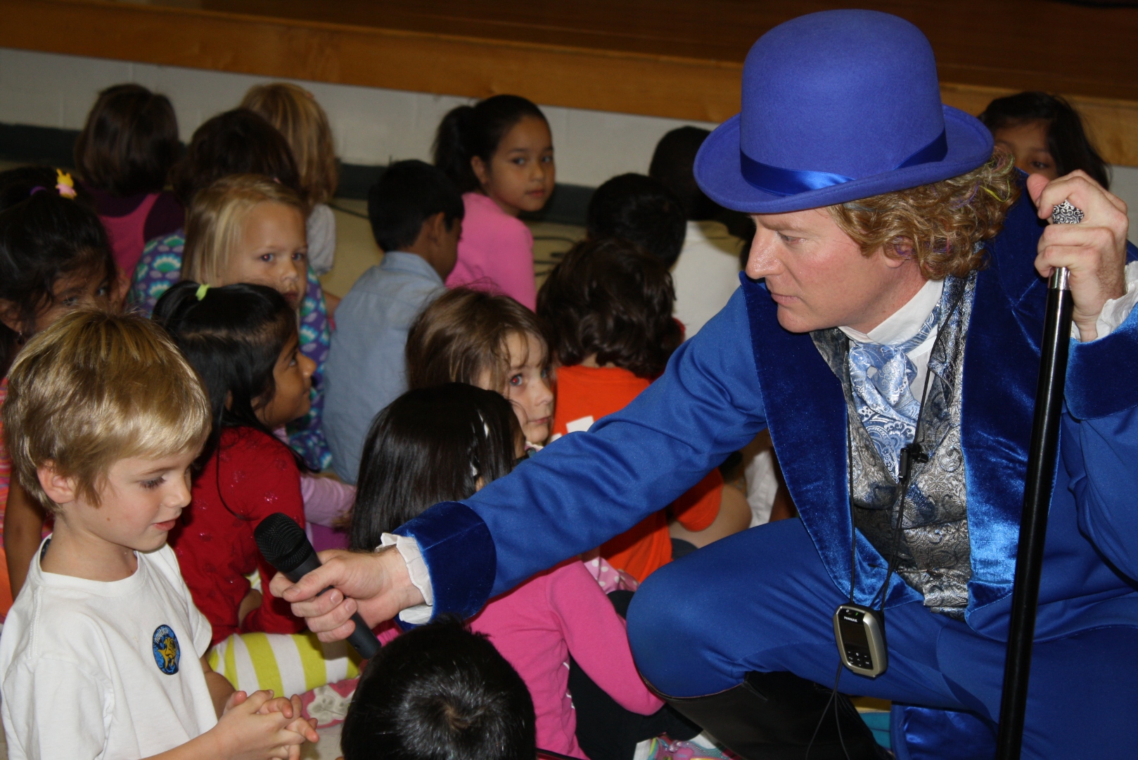 Wylde Scott in his blue costume and top hat kneeling to hand the microphone to a young student surrounded by captivated children during a school performance