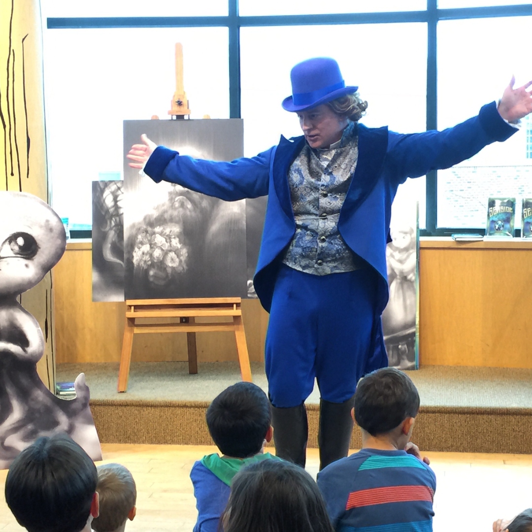 Wylde Scott performing live for children in his signature royal-blue suit and matching top hat, arms thrown wide mid-story.